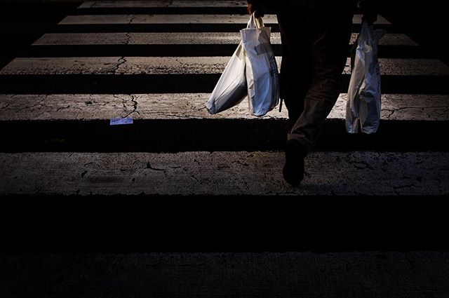 A person carries shopping bags while crossing a zebra crossing in a street, casting shadows with high contrast.
