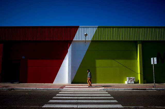 A man walks across a zebra crossing against a vibrant red, white, and green building under a clear blue sky.