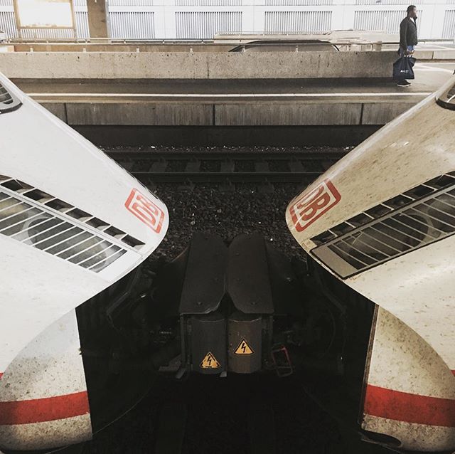 Two modern trains sit on the tracks at a station while a person walks on the platform.