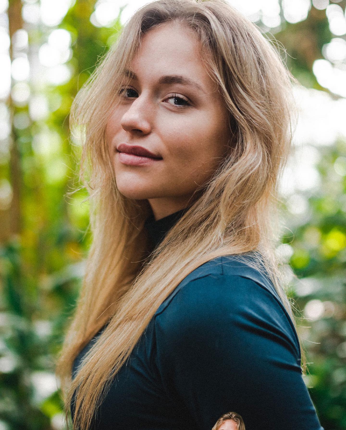 A blonde woman poses outdoors in a natural setting with soft, diffused lighting.