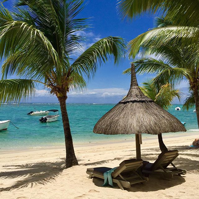 A serene beach scene with palm trees, beach chairs, and boats on tranquil turquoise water under a clear sky.