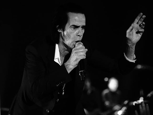Black and white close-up of Nick Cave singing into a microphone during a live performance.