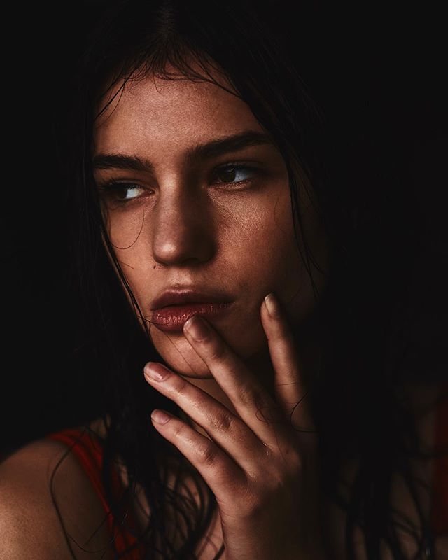 A brunette woman gazes intensely with her hand near her face in a moody, close-up portrait.