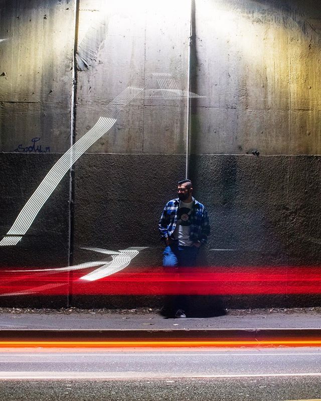 A man leans against a concrete wall at night with streaks of light creating an urban scene.