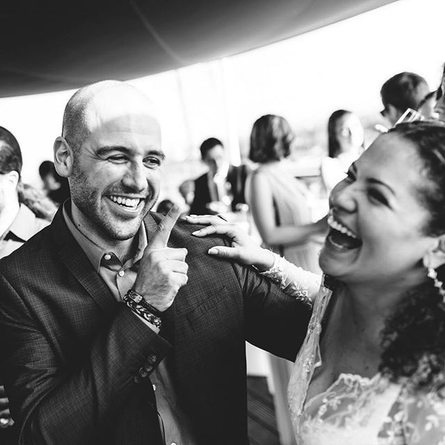 A smiling couple laughs together at an outdoor wedding celebration, captured in a candid black and white photograph.