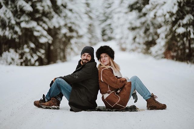 A couple in winter clothing sit back-to-back in the snow on a forest path.