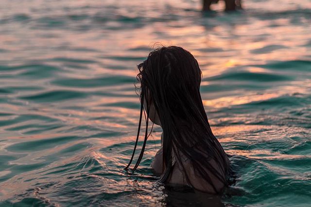 A woman with long dark hair stands in the ocean, enjoying a peaceful moment at the beach during sunset.