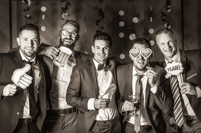 A group of groomsmen celebrate at a wedding photo booth with props and suits in a black and white image.