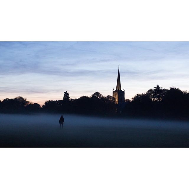 A lone figure stands in a foggy field at dusk with a church spire in the background, creating a mysterious scene.