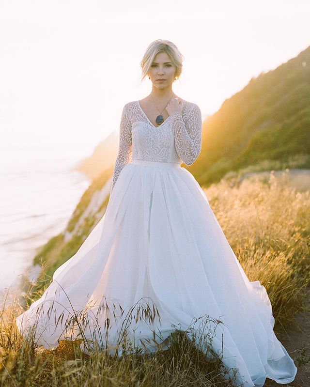 A bride in a white lace dress stands on a cliffside at sunset.