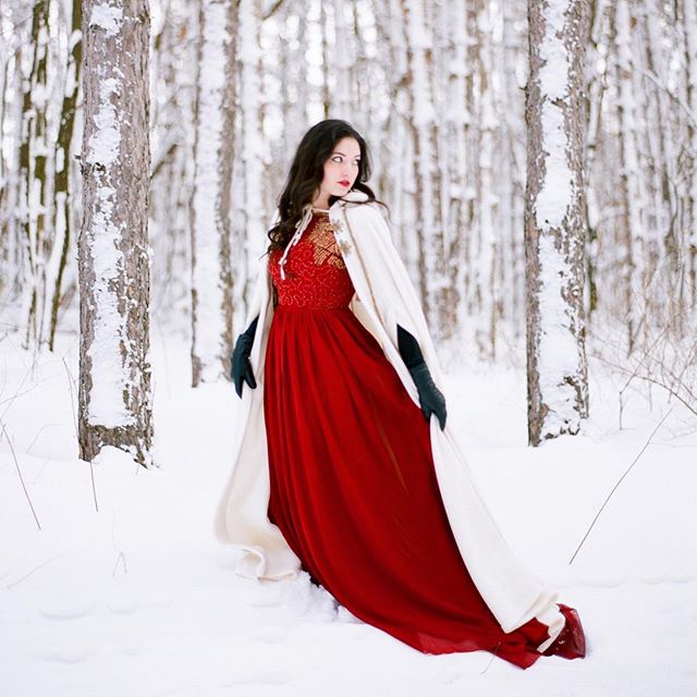 A woman in a red dress and cape stands in a snowy forest, embodying a winter fairytale scene.