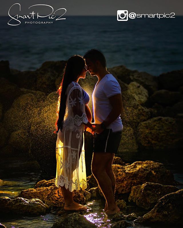 A pregnant woman and a man share an intimate moment on a rocky beach at dusk, illuminated by a warm backlight.
