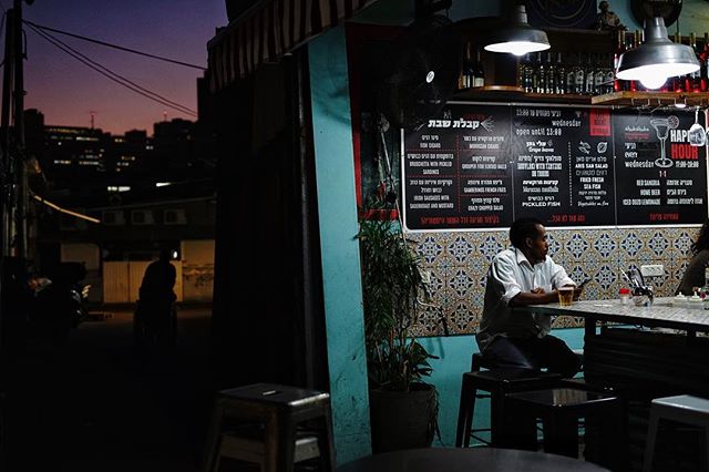 A man sits at a bar at night, reading a menu with liquor bottles on the shelf behind.