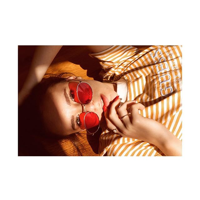 A woman with red sunglasses and a striped top lies on a wooden surface in a sunlit, stylish shot.