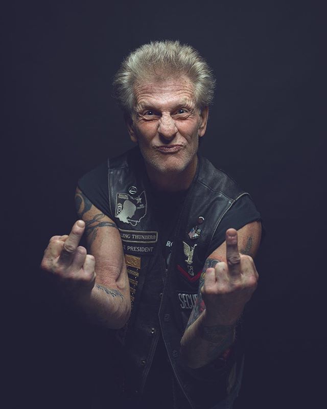 An older biker confidently makes a rude hand gesture in a studio portrait, tattoos visible.
