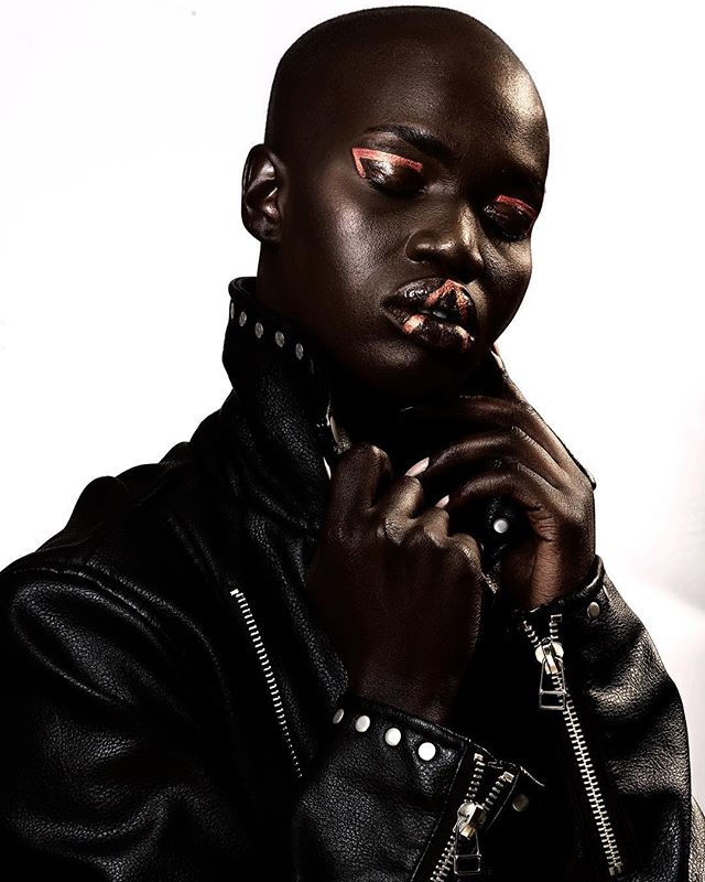 A bald person with creative makeup in a black leather jacket poses in a studio.