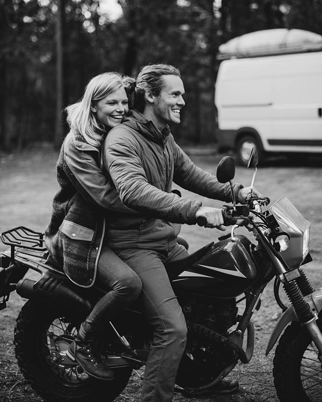 A smiling couple rides a motorcycle in a forest with a van in the background.
