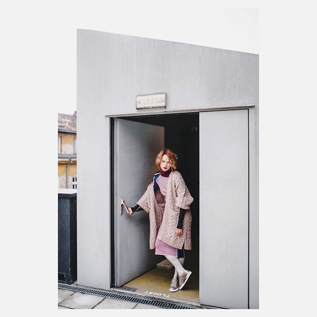 A woman in a pink outfit and beige cardigan poses in a doorway for a fashion editorial.