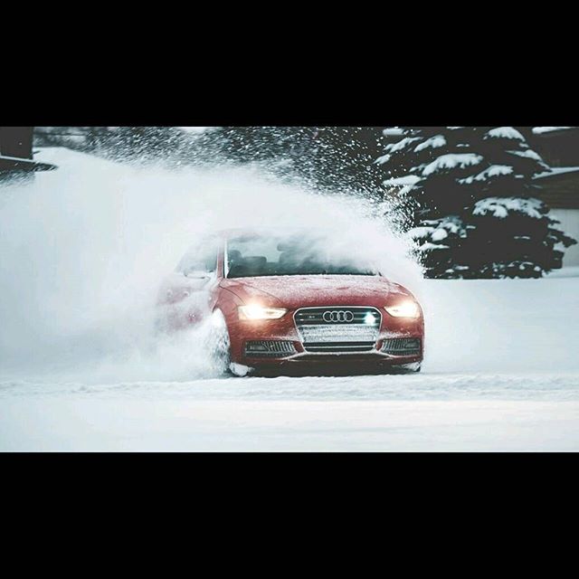 A red Audi A4 drives through the snow, kicking up a cloud of white powder in a winter landscape.