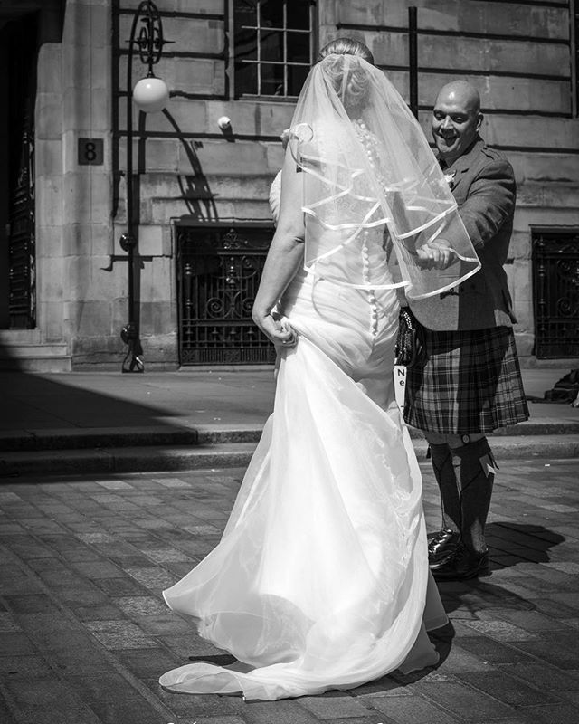 A bride in a white dress and veil stands with a groom in a kilt on a city street.