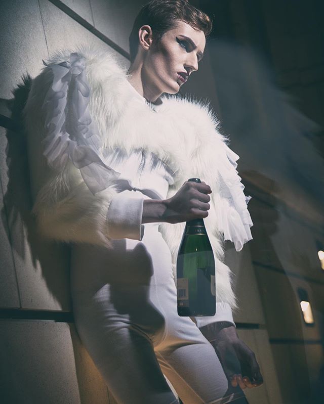 A man in white attire stands with a champagne bottle in an editorial fashion shot.