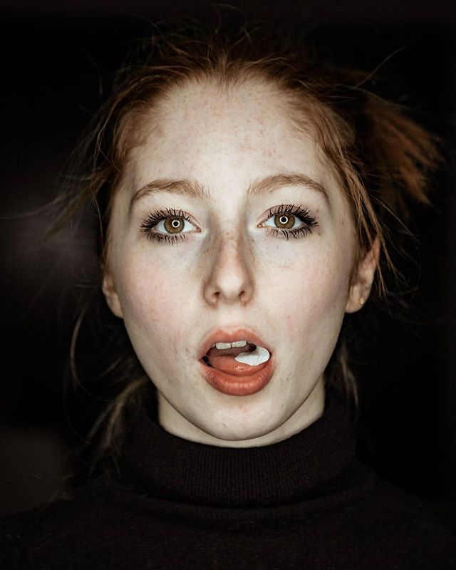 A striking redhead woman poses in a close-up studio portrait, adding a playful touch with chewing gum.