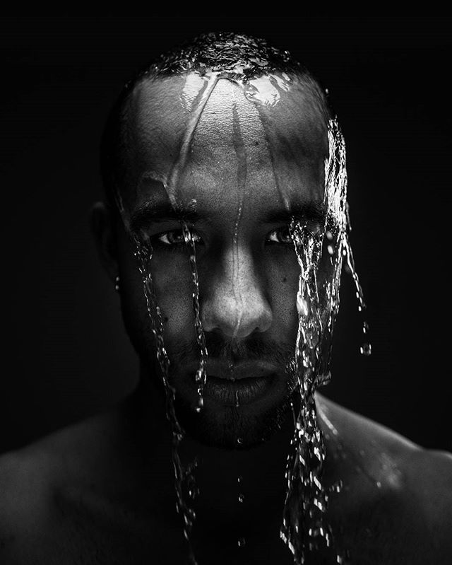 A dramatic black and white portrait of a man with water cascading down his face against a dark background.