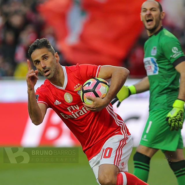 A soccer player celebrates holding the ball during a soccer game with goalkeeper in the background.