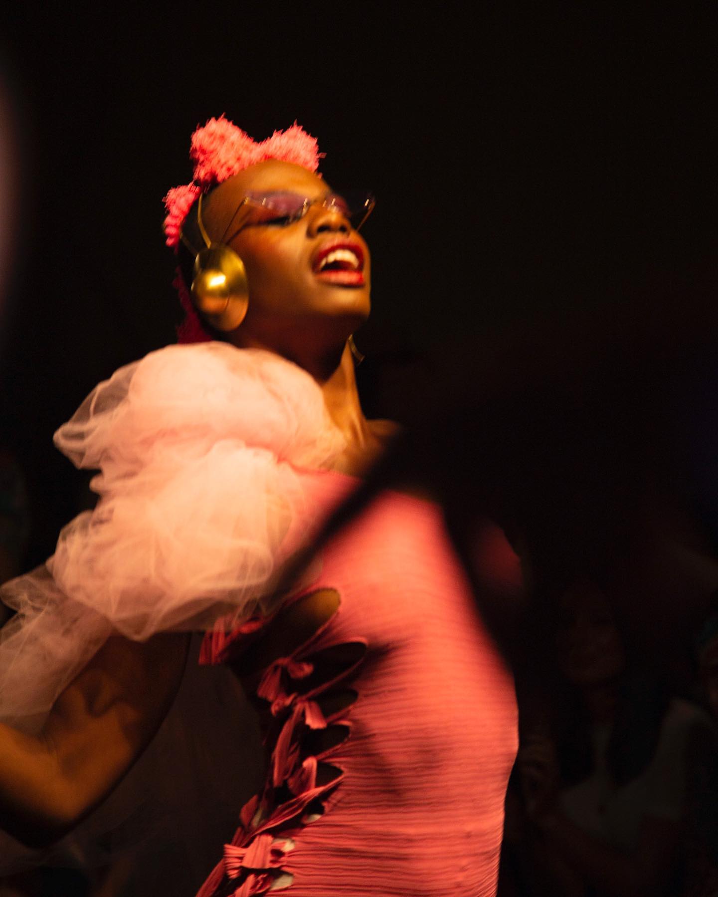 A vibrant performer in a pink outfit poses against a dark backdrop, exuding energy and confidence.