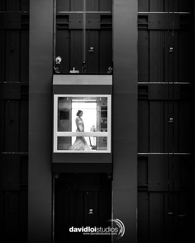 A bride in her wedding dress stands inside a unique elevator, framed by the architecture in a striking monochrome shot.