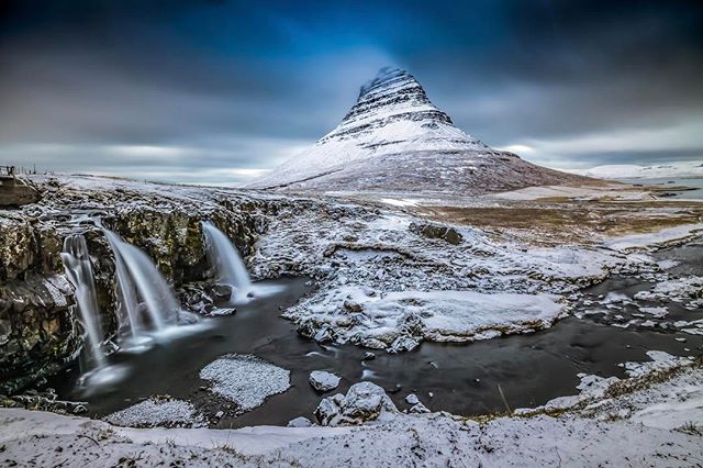 A scenic winter landscape view of Kirkjufell mountain and waterfalls in Iceland under a cloudy sky.