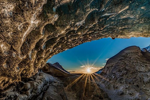 A scenic landscape showcases the interior of an ice cave, with sunlight streaming through the opening and illuminating the mountains beyond.