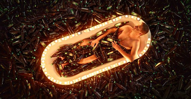 Man relaxes in a bathtub filled with beer bottles, illuminated by warm, glowing lights.