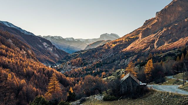 A scenic view of a valley with autumn-colored trees, mountains, and a rustic cabin nestled near a winding road.