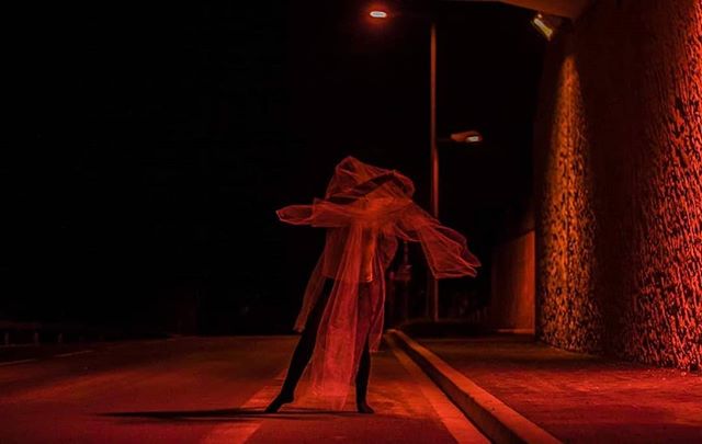 A solitary figure shrouded in red light stands on an empty road at night, creating a mysterious and moody atmosphere.