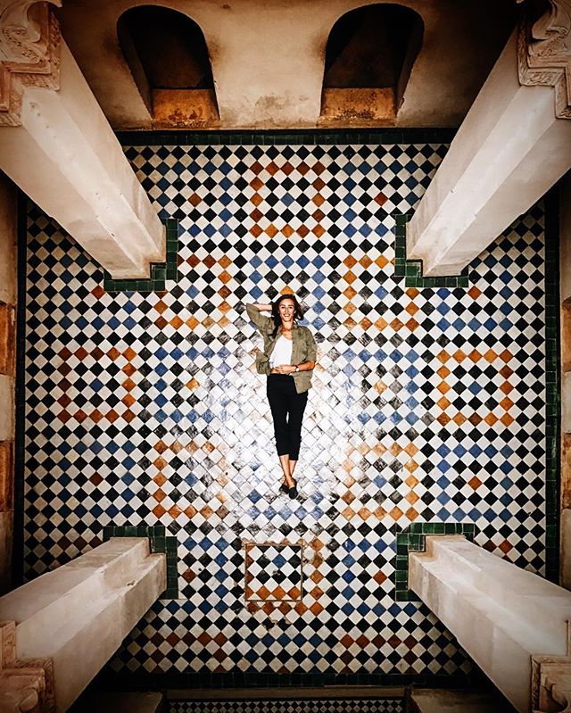 A woman lies on a patterned tile floor in an architectural space with overhead beams.