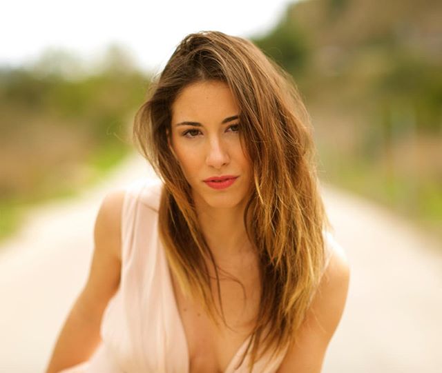 A brunette woman poses outdoors, looking at the camera with soft lighting in a close-up portrait.