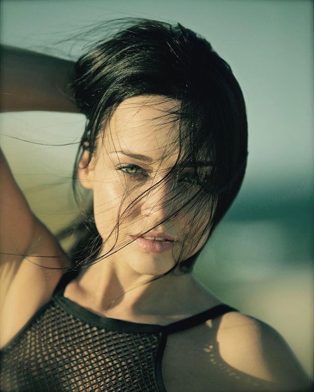 A brunette woman poses on a beach, hair blowing in the wind, for a sensual portrait.