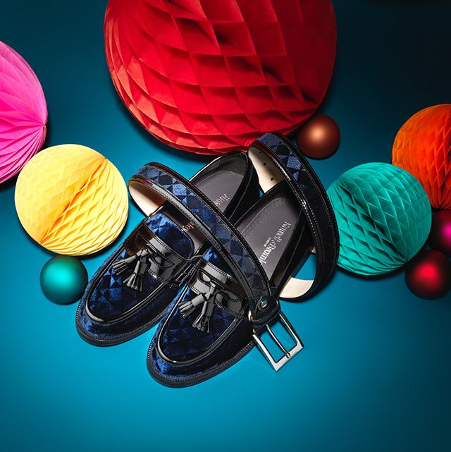 A stylish pair of blue velvet loafers and a black patterned belt are displayed against a vibrant teal background, surrounded by colorful decorations.