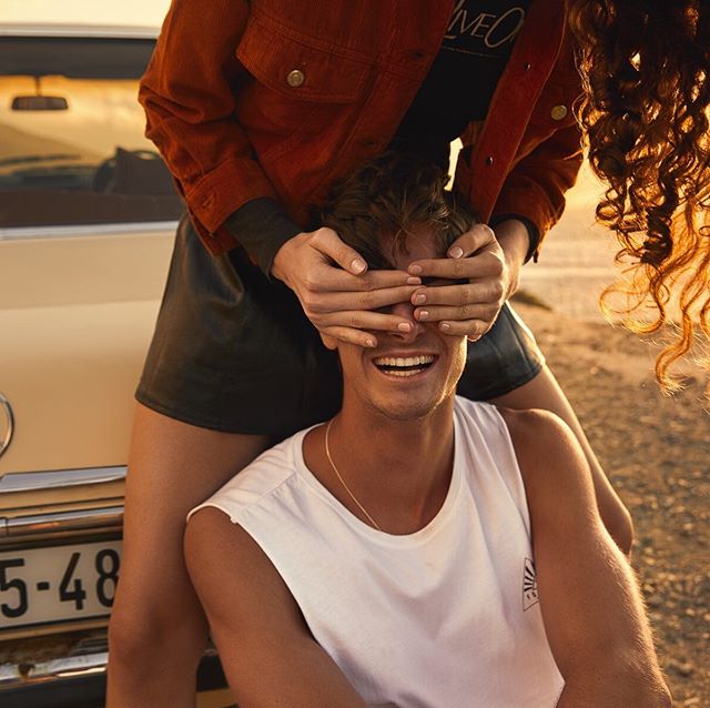 A smiling man sits as a woman playfully covers his eyes near a vintage car at the beach.