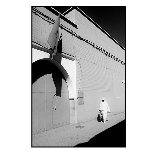 A person in white religious clothing walks past a building on a street in a high-contrast black and white image.