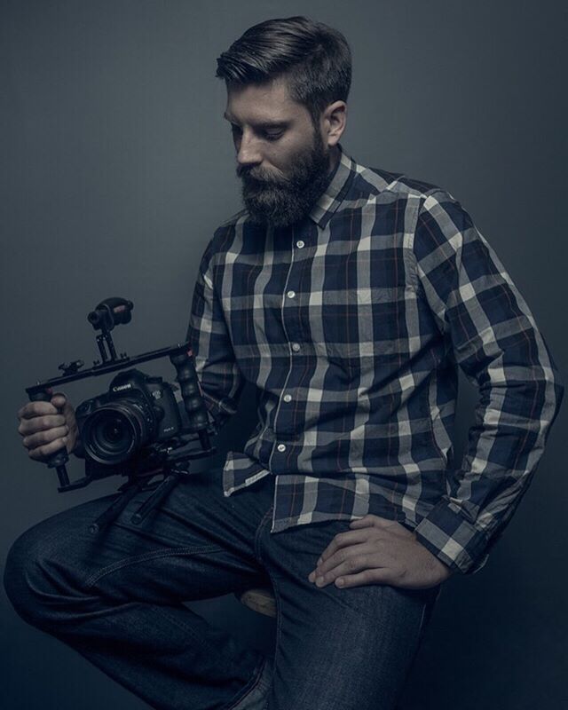 A bearded man in a plaid shirt is sitting and holding a Canon camera.