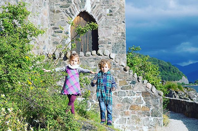 Two young children stand outside a stone castle on a sunny day, exploring and enjoying a family trip.