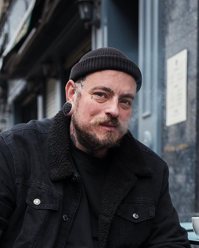 A tattooed man with a beanie smoking, wearing a denim jacket in an urban setting, looking at the camera.