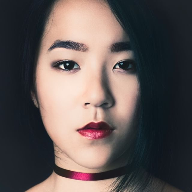 Close-up portrait of a serious Asian woman with dark hair and red lipstick against a dark background.