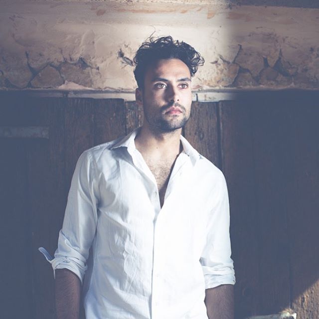 A man in a white shirt stands indoors looking thoughtfully into the distance.