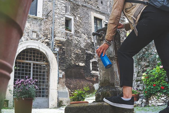 A person filling a blue water bottle at a stone fountain in a european town.