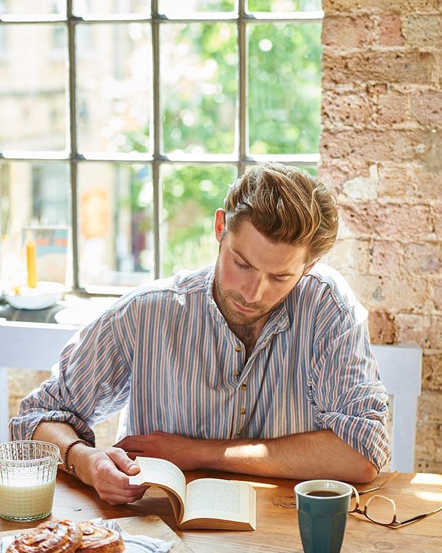 A man relaxes and reads a book at a table with coffee and pastries in a sunlit room.