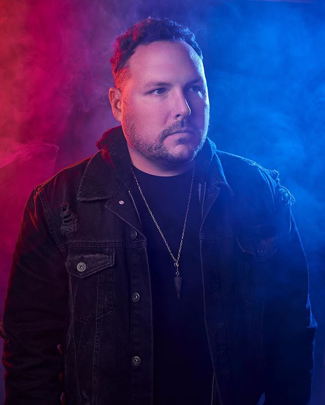 A portrait of a man in a denim jacket against a blue and red smoky background.