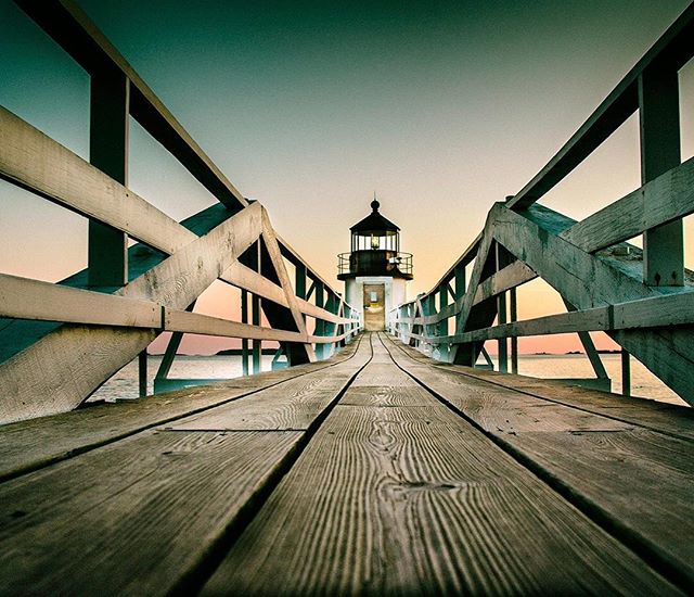 A serene lighthouse stands at the end of a wooden pier against a tranquil sky and ocean backdrop.